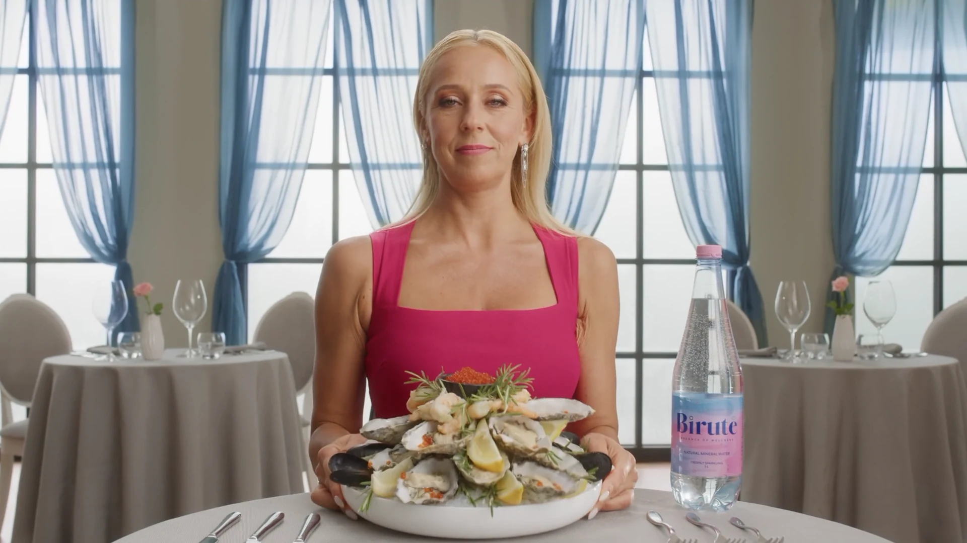 Birutės reklamos kadras restorane: aktorė su didžiule jūros gėrybių porcija ir mineralinio vandens buteliu.
